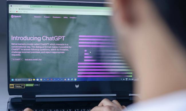 A person sits in front of a laptop. On the laptop screen is the home page for OpenAI's ChatGPT artificial intelligence chatbot.